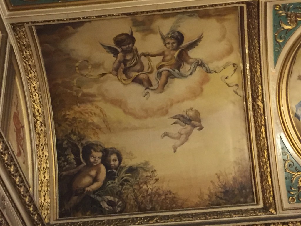 A large painting on the lobby ceiling in the Criterion Theatre. At the top, a pair of cherubs with wings sit on a large cloud together, each holding a separate length of coloured ribbon, and in each case the other end of their ribbon is floating around in the air next to them. Another cherub is flying through the air below them, and 2 more are sitting amongst the grass in the bottom left corner, one of them holding a lute instrument.
