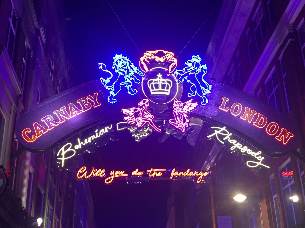 Under the night sky, a dark blue arched sign has the words Carnaby and London on each side of it in orange capitals. Just below the arch, again on each side, are the words Bohemian and Rhapsody in lit-up neon yellow curly script lettering. In the centre of the arch is the Queen crest lit up in various colours. An orange crab sits on a yellow letter Q with a yellow crown in the middle of it. 2 blue lions, one on each side, are holding up the letter Q with their front legs while standing on their back legs. 2 pink fairies are seated either side of the Q at the bottom. In the background another sign in curly orange letters says Will you do the fandango?