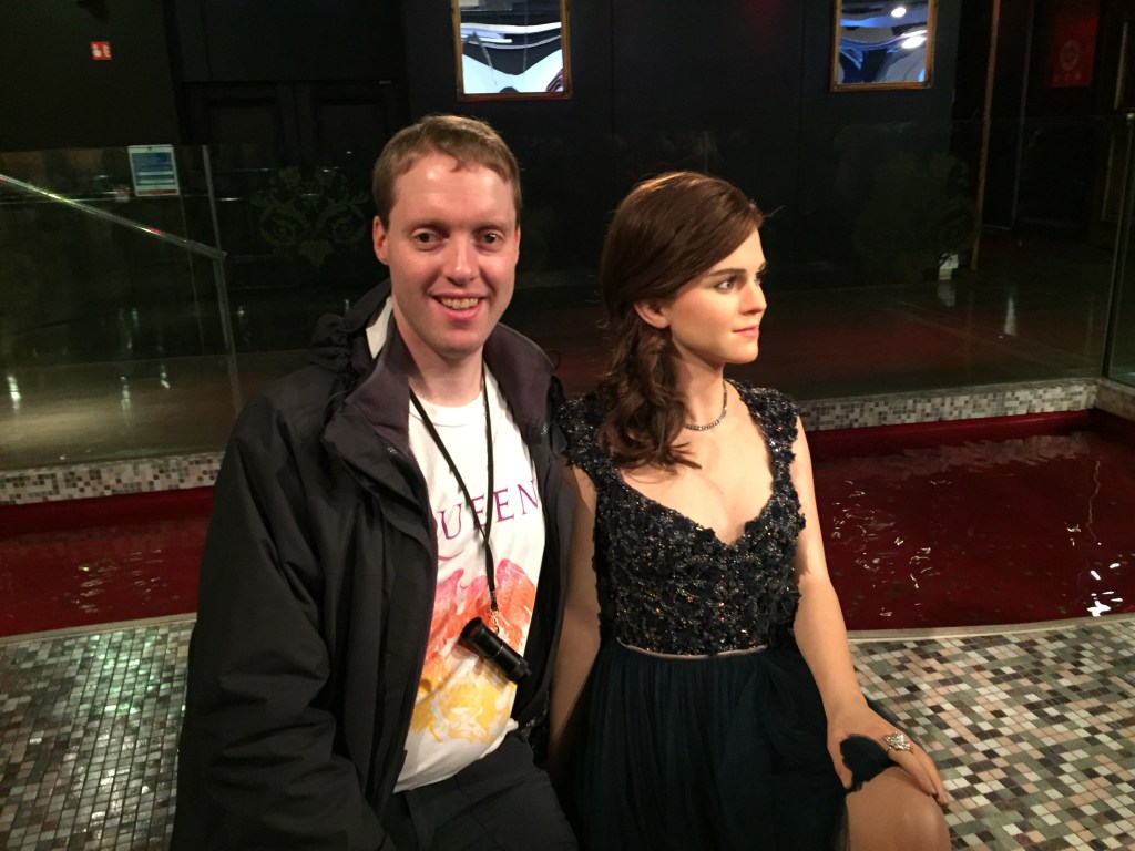 Glen smiling as he sits with a waxwork of Emma Watson, who is wearing a sparking black dress.