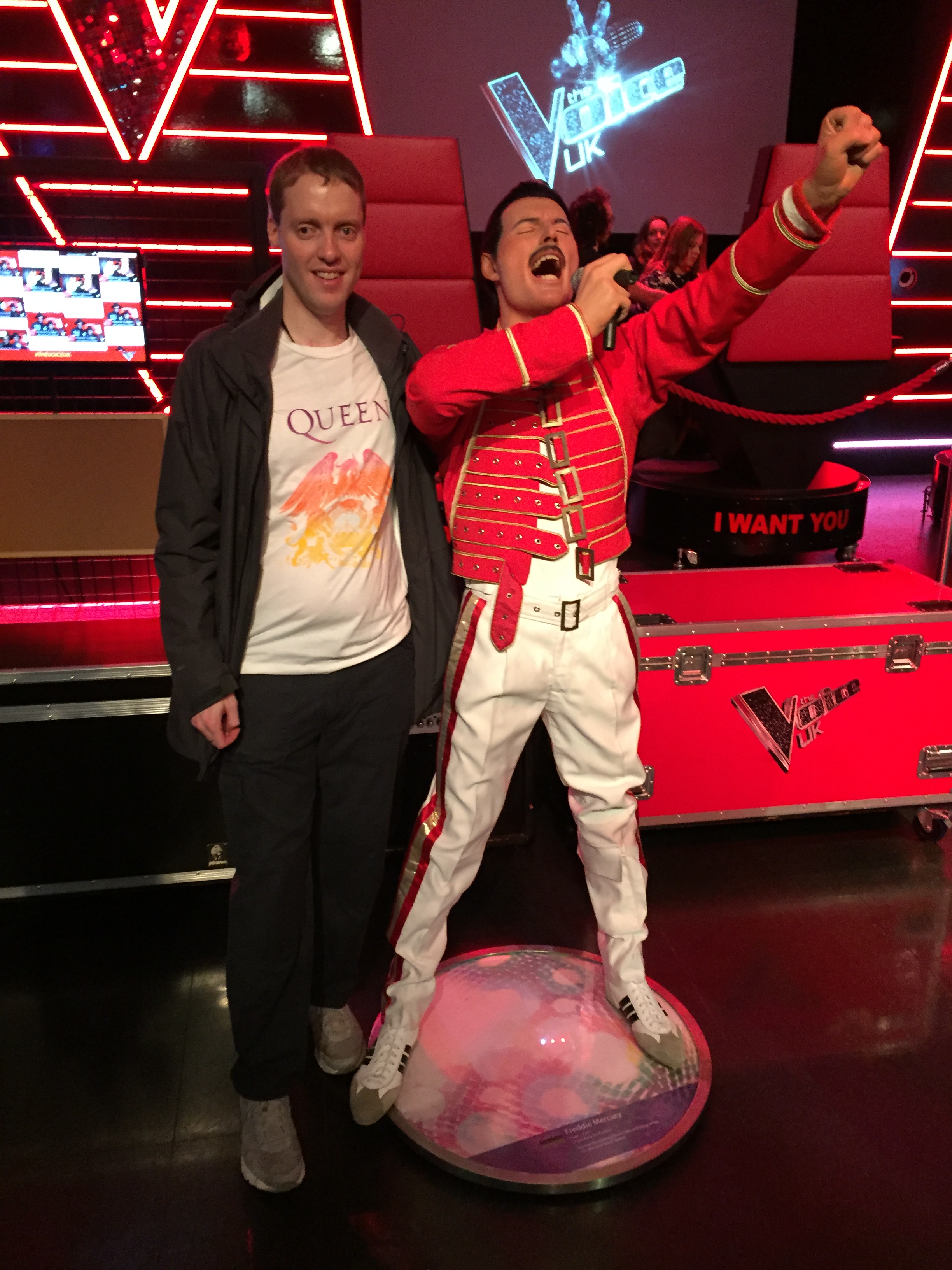 Glen smiling and wearing a white t-shirt featuring the colourful crest for the band Queen, as he stands next to a waxwork of Freddie Mercury. Freddie has a red jacket and white trousers on, and is punching the air with his left hand as he sings loudly into the microphone held in his right hand.