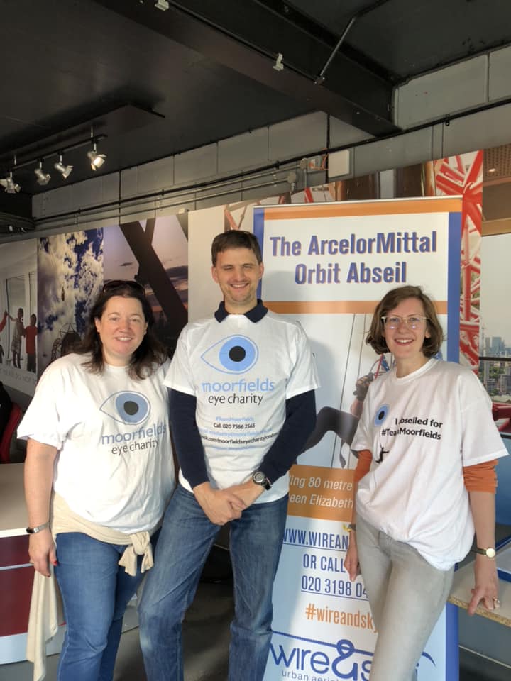 James Buller posing with another female abseiler in their Moorfields Eye Charity abseil t-shirts, along with a lady from Moorfields Eye Charity, wearing a regular white t-shirt with the charity's logo on the front, a blue circle with a black pupil in the centre.