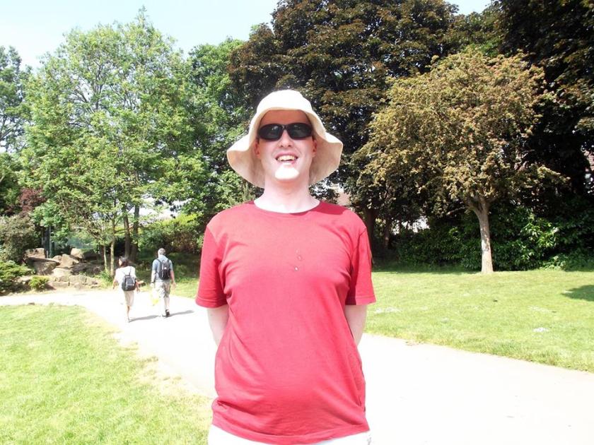 Glen smiling while standing in Lloyd Park in the sunshine, wearing a sunhat, sunglasses and a red t-shirt.