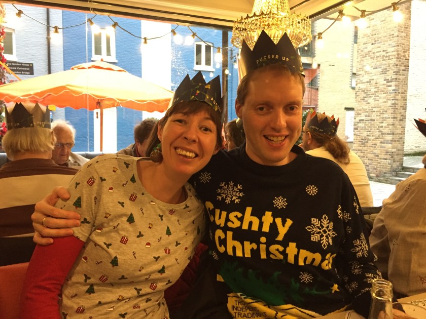Glen with his arm around Jessica as they sit together and smile at the camera. Both are wearing Christmas hats from their Christmas crackers. Glen is wearing an Only Fools And Horses Christmas jumper, with large yellow letters saying Cushty Christmas above the yellow 3 wheeled van owned by the Trotters in the TV show, surrounded by snowflakes. Jess is wearing a white top covered in little Christmas trees, holly leaves, etc, with red sleeves.