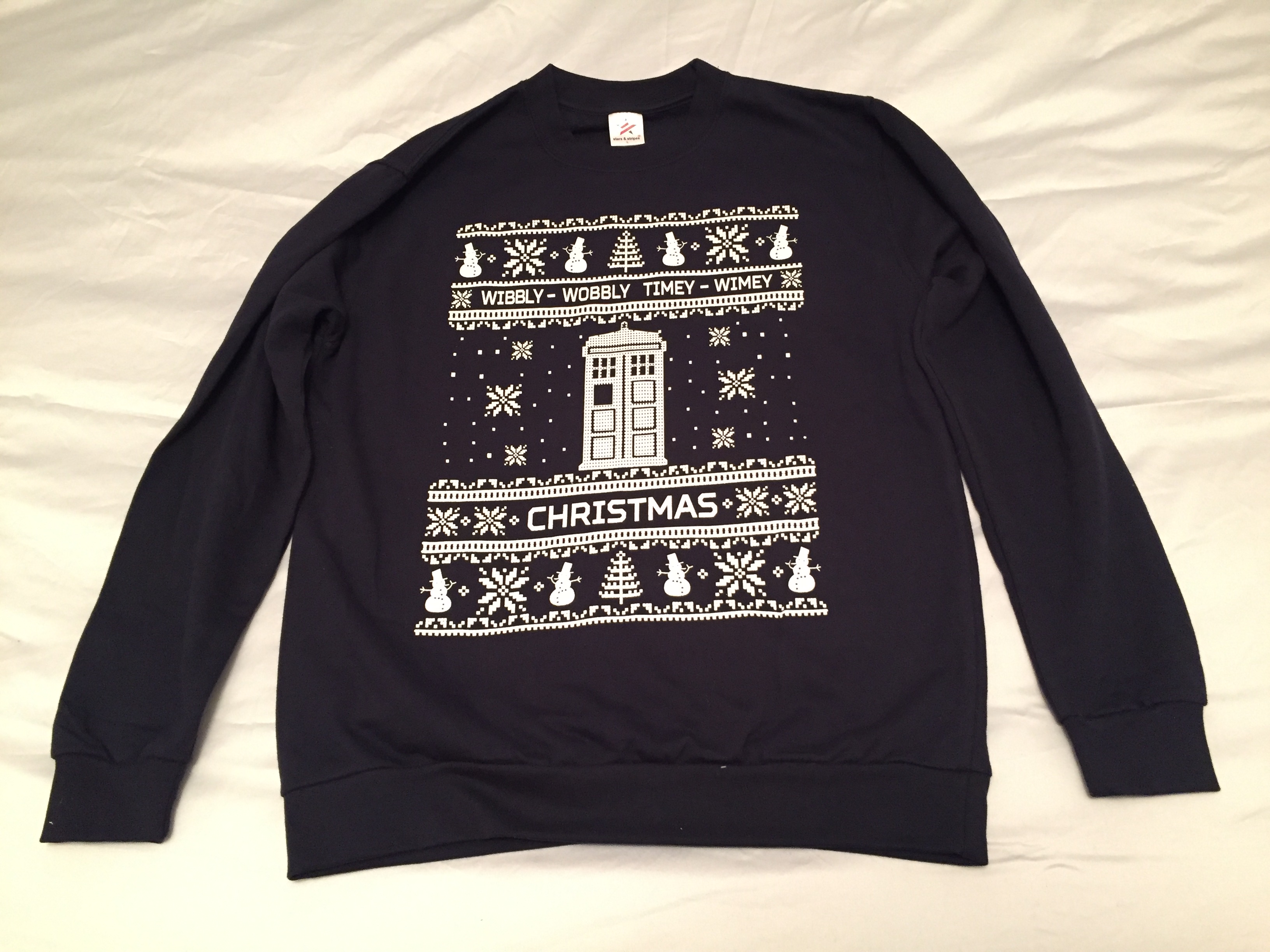 A dark navy jumper with a white design in the centre. The centre shows a large white Tardis with snowflakes falling around it. Above it are the words Wibbly Wobbly Timey Wimey, and below is the word Christmas. At the very top and bottom is a row of snowmen, snowflakes and Christmas trees.