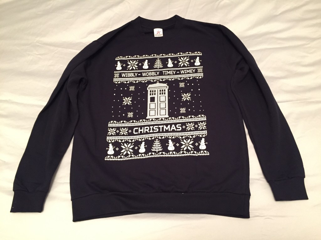 A dark navy jumper with a white design in the centre. The centre shows a large white Tardis with snowflakes falling around it. Above it are the words Wibbly Wobbly Timey Wimey, and below is the word Christmas. At the very top and bottom is a row of snowmen, snowflakes and Christmas trees.