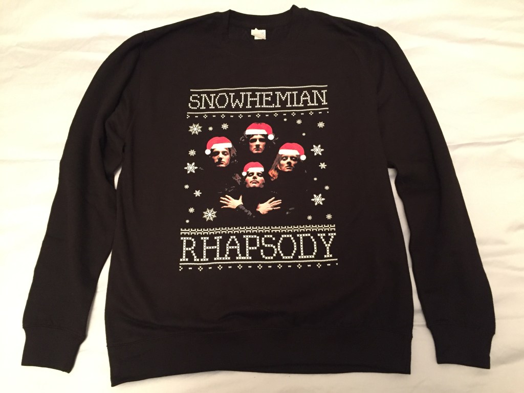 Black jumper with an image of the band Queen in the centre. The 4 members of the band are shown as headshots, with Freddie in the front, Brian at the back, John on the left and Roger on the right, in the pose from their Bohemian Rhapsody video. But here they all have red and white Santa hats superimposed on their heads. The image of the band is surrounded by snowflakes, and it all sits between the words Snowhemian Rhapsody, printed in large white letters.