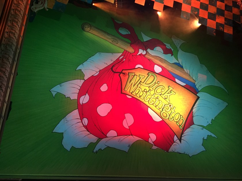 Stage curtain for the Dick Whittington pantomime, showing a large red bag with white spots strapped to the end of a wooden pole, like the one Dick carries his belongings in over his shoulder during the show. A large gold label attached to the bag says Dick Whittington in large letters.