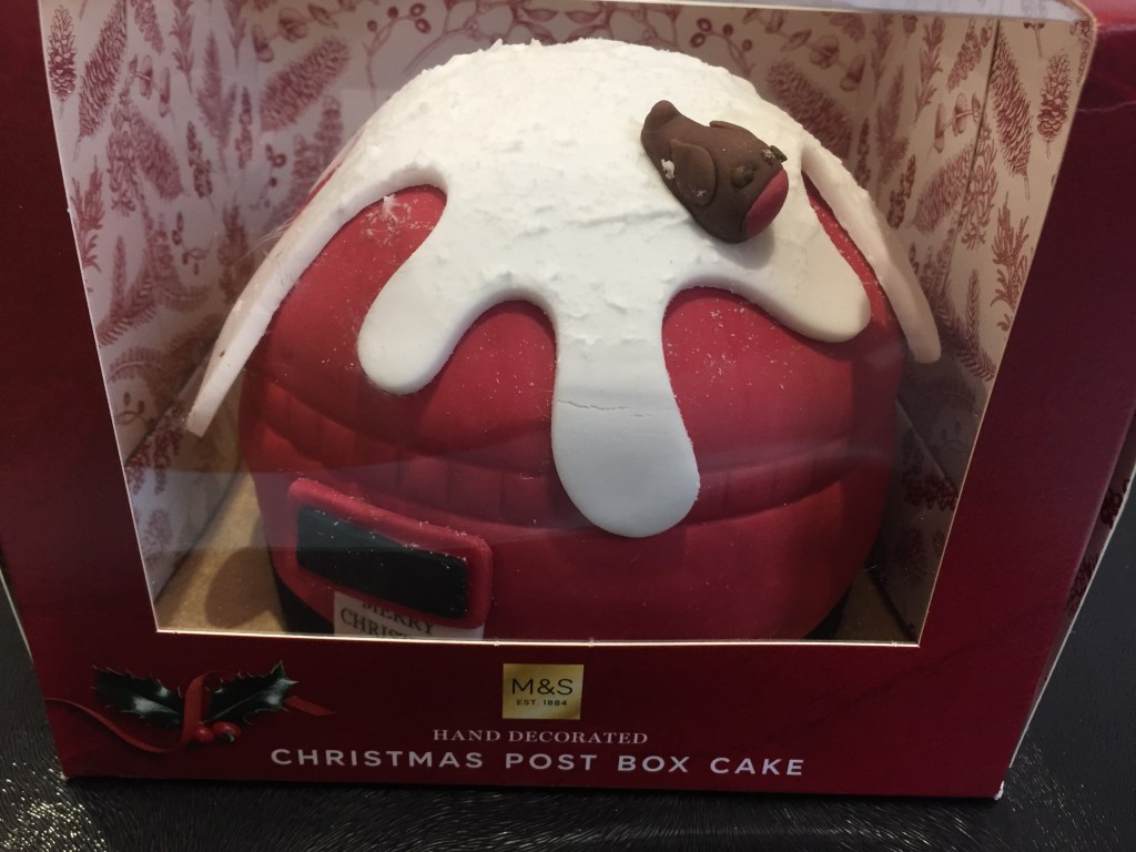Marks & Spencer hand decorated Christmas Post Box Cake. The large cake looks like the top round section of a red post box, with the black letter box opening on the side, and a layer of white snow covering the top, with a robin perched on it.