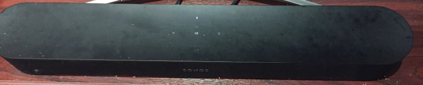 Sonos Sound Bar, a long narrow speaker with curved ends, and a few small buttons on the top to adjust the volume and activate the Alexa voice assistant.