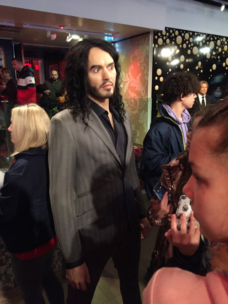Madame Tussauds waxwork of Russell Brand wearing a grey suit with faint vertical stripes in alternating shades, and a black shirt underneath.