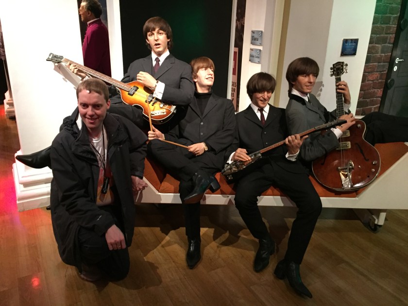 Glen kneeling and smiling towards the camera next to a long couch on which are waxworks of The Beatles. Ringo Starr is holding his drumsticks, while the other 3 are playing their guitars.