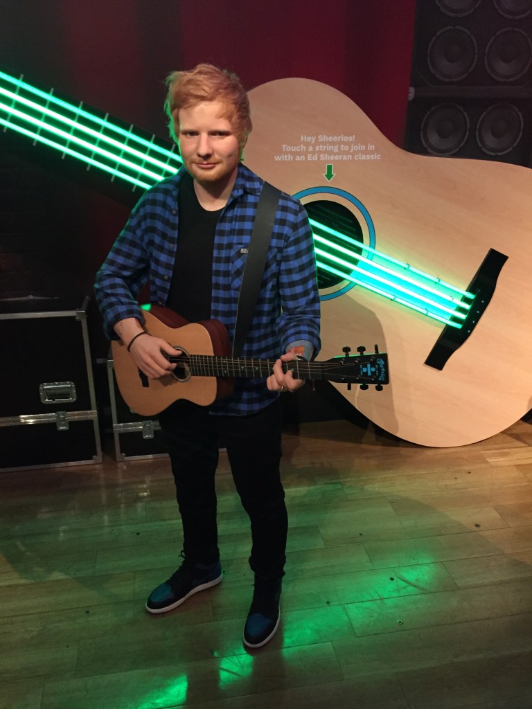 Waxwork of Ed Sheeran at Madame Tussauds. He is wearing a blue and black checked shirt over a black t-shirt, and black trousers, and is playing his guitar. A huge guitar on the wall behind him has 4 long strings that are glowing green. Text above it says Hey Sheerios, touch a string to join in with an Ed Sheeran classic.