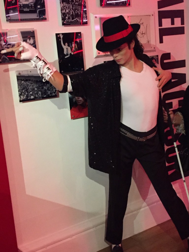 Waxwork of Michael Jackson at Madame Tussauds. He is striking a pose while wearing a black hat with a red stripe around it, a black jacket and trousers, and a white t-shirt.