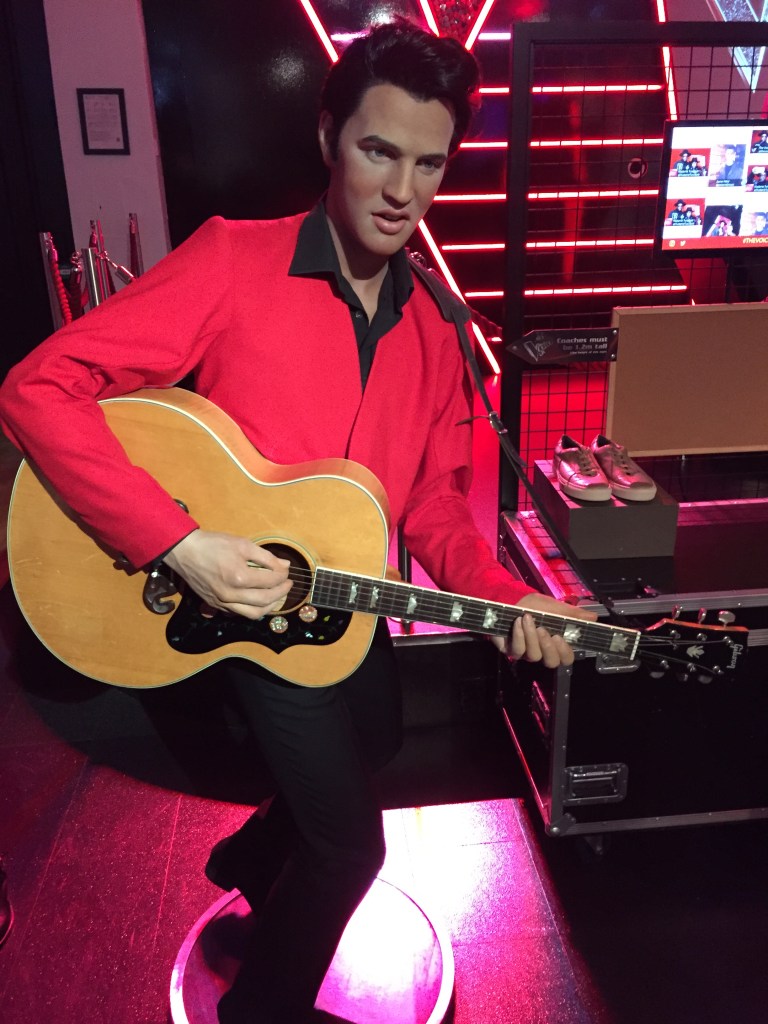 Waxwork of Elvis Presley at Madame Tussauds. He's wearing a red jacket with a black collar, and black trousers, while playing the guitar.