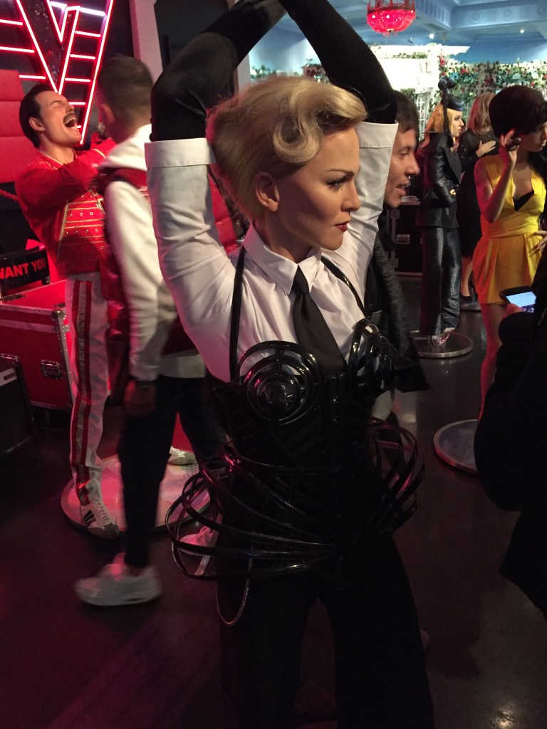 Waxwork of Madonna at Madame Tussauds. She has her arms stretched above her head. The top part of her outfit has a white shirt and a black tie, but from her breasts downwards the outfit is black. Round plastic objects sit over her breasts on the outside of her outfit, and more round objects fan out from her hips on either side.