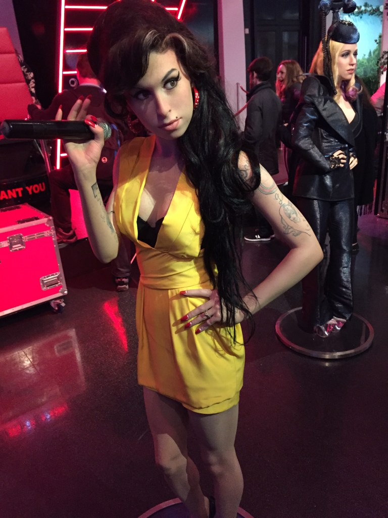 Madame Tussauds waxwork of Amy Winehouse, who is wearing a yellow dress, holding a microphone in her right hand, and has her left hand on her hip as she looks to her left.