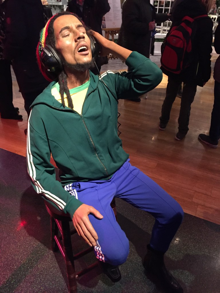 Waxwork of Bob Marley at Madame Tussauds. He is sitting with his eyes closed as he enjoys the music on his headphones, while wearing a green jacket with white stripes along the side of the sleeves, and blue trousers with shite stripes down the side of the legs.