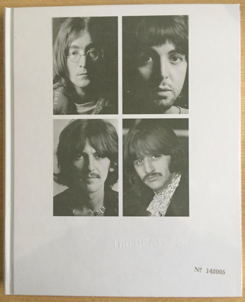 Cover for the CD box set of The White Album by The Beatles. The set is a large white book, overlaid with a transparent sleeve, which is dominated by 4 portrait photos, 1 of each of the band members. In the bottom-right corner, on the book itself beneath the sleeve, is a stamp saying number 142005. To the left and slightly above this on the book cover is embossed white text, which is almost invisible, saying The Beatles.