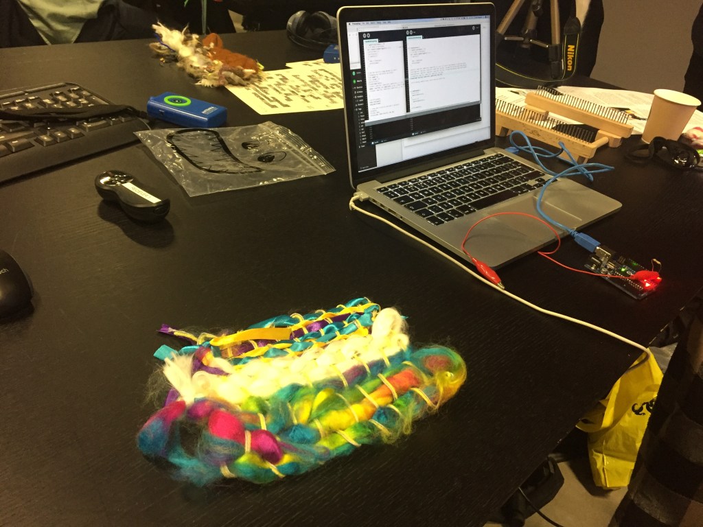 Our colourful woven creation, made of blue and yellow ribbons, white wool and rainbow coloured wool, on the desk next to a laptop, into which is plugged a small device like a Raspberry Pi, to be connected to our craftwork.