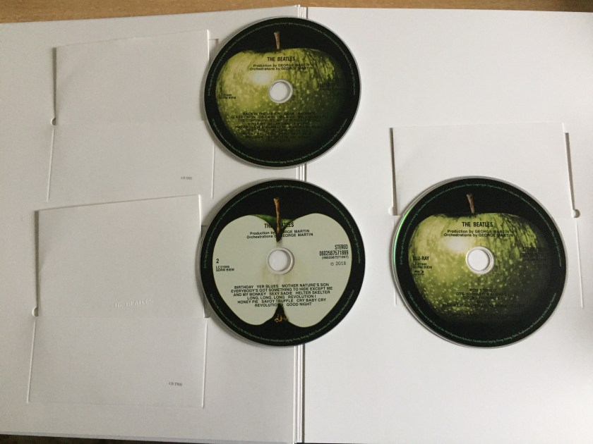 The inside front cover of the large book for The White Album boxset, showing the first 2 CDs and Blu-ray disc, and the white sleeves they came out of. The discs are each filled with a large image of an apple, which is whole on CD 1 and the Blu-ray, and cut in half on CD2 to reveal the white centre. The track listing on each disc is on top in black text.