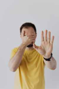 A man covering his eyes with his right hand, while holding up his left hand, palm facing us, as a signal to stop.