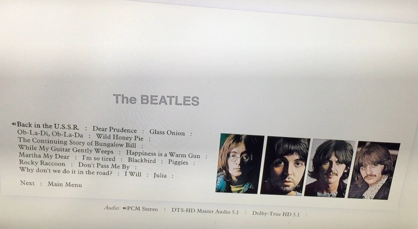 Album tracks menu on the Blu-ray disc for the White Album box set. On a white background, the tracks for the first half of the album are in black on the left, while on the right are 4 portrait photos of the band members. Audio options for stereo and 5.1 sound are at the bottom of the screen, and just above the track listing is larger grey text saying The Beatles.