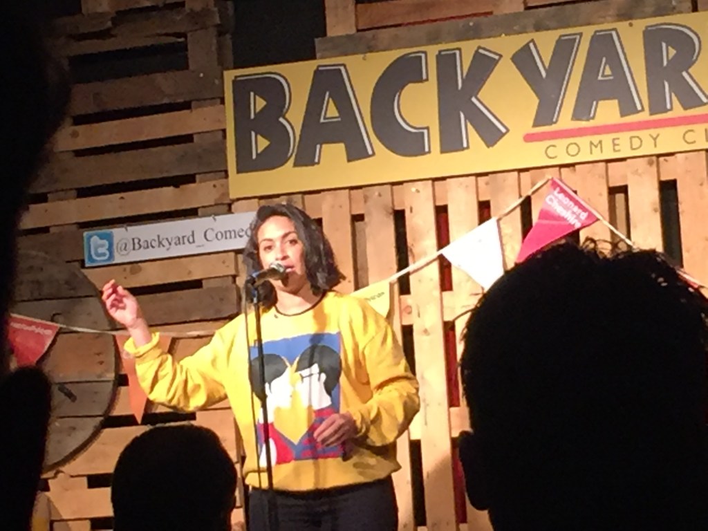 Emily Lloyd-Saini on stage at the Backyard Comedy Club. She is wearing a yellow jumper, on the front of which is artwork of a man and woman looking at each other. both with dark hair and the man wearing glasses.