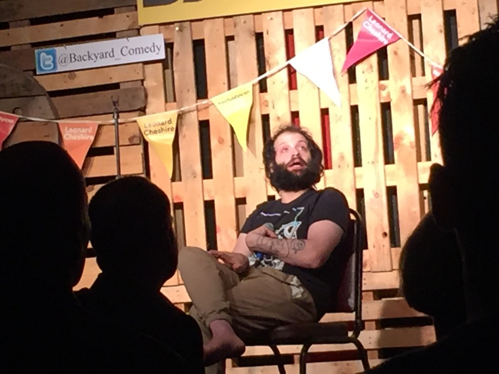 Tim Renkow, sitting on a chair on the Backyard Comedy Club stage as performs his set. He is wearing a black t-shirt, the white pattern of which is obscured by his left arm across his chest, which has a tattoo covering his forearm.