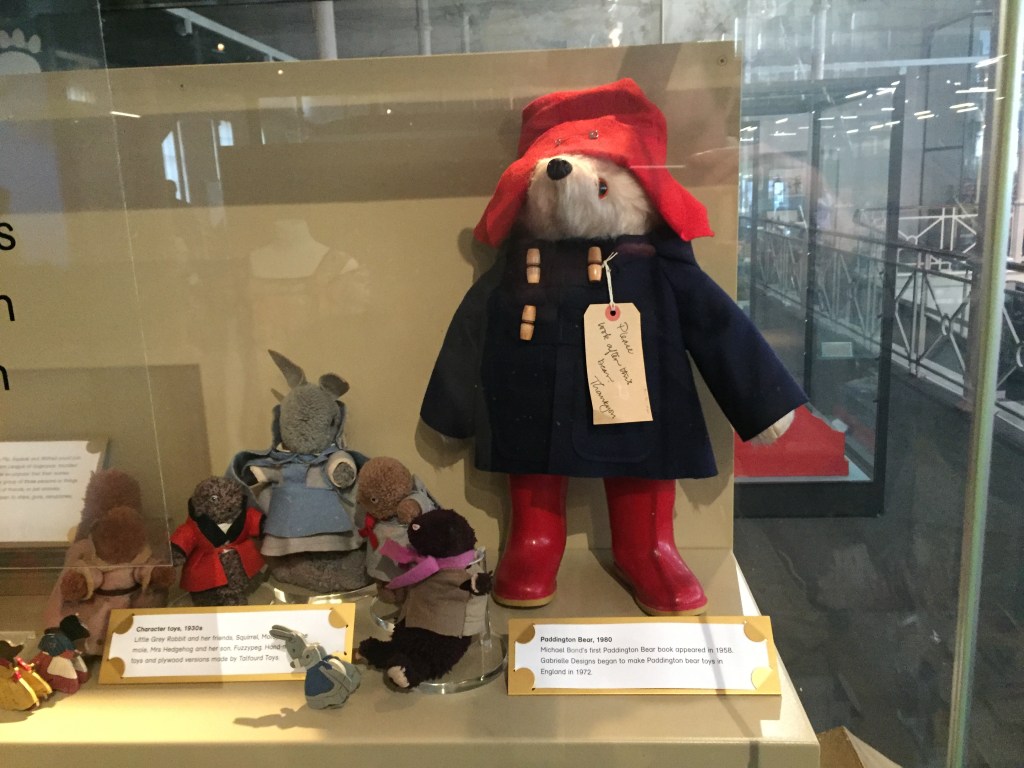 A large stuffed Paddington Bear toy, wearing a red hat, navy coat and red wellies, with a baggage label tied to one of his coat buttons that reads Please look after this bear, thank you. Next to him is a much smaller stuffed toy of Little Grey Rabbit, with her even smaller rabbit daughter and her squirrel and hedgehog friends.