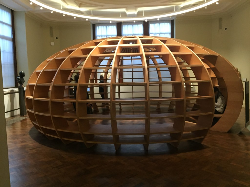 A large globe-shaped wooden structure, like a squashed ball, filling the centre of the room. The outer walls are a wooden grid all the way around, effectively creating lots of little shelves. A tall opening allows people to walk inside and sit on the circular bench round the inner edge of the structure.