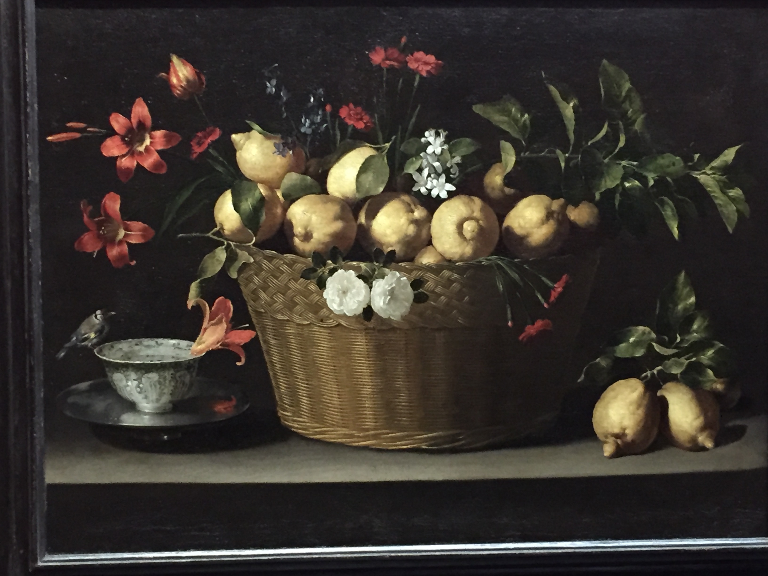 Still life painting. In the centre is a large woven basket filled with fresh lemons, along with sprigs of lemon blossom, red carnations, blue delphiniums, two white roses, day lilies and a tulip. in the bottom left is a silver bowl filled with water, on a blue-and-white porcelain bowl. A goldfinch perches on the lip of the bowl, and a day lily floats in the water.