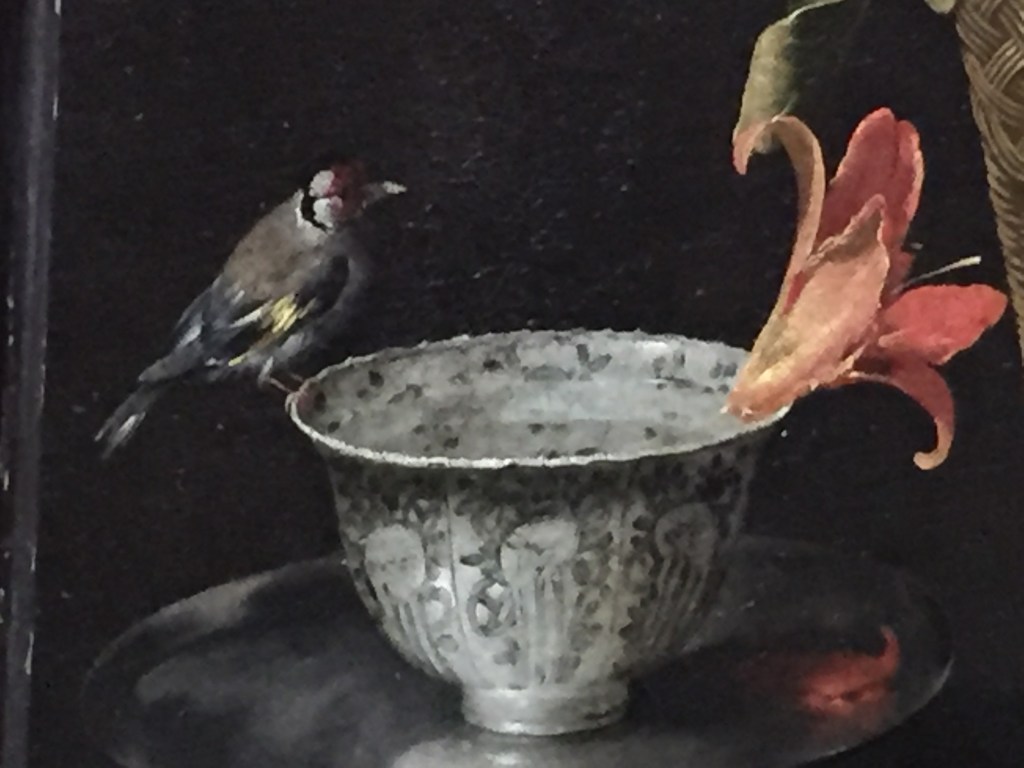 Close-up of the painting Still Life Iin A Wicket Basket, focusing on a goldfinch perched on a decorative silver bowl filled with water, opposite a day lily floating in it on the other side.