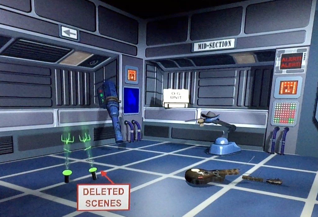 The extras DVD menu for Red Dwarf series 6. The screen shows a room with metal walls, some panels of buttons, and a floor made of blue squares with white borders. Moving around the screen allows you to select various items to access different features, including a broken guitar, a skutter robot and a bazookoid gun.