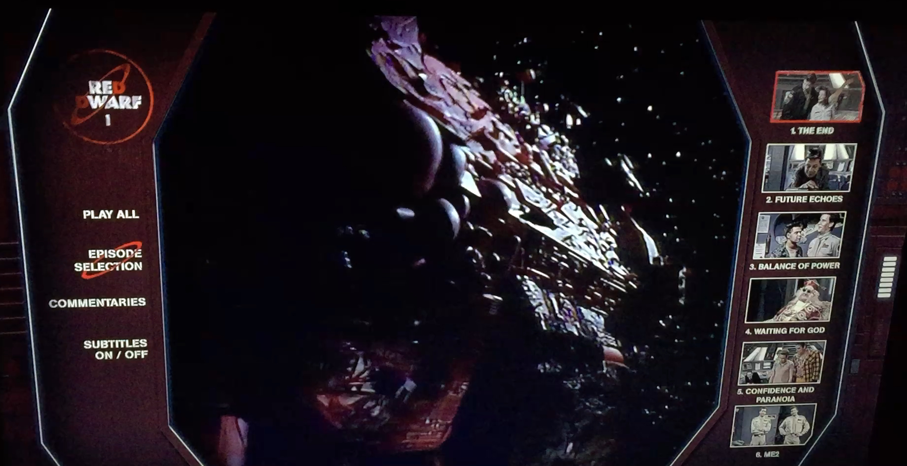 Menu for the Blu-ray of Red Dwarf series 1. In the centre of the screen is an image of the huge Red Dwarf spaceship flying through space. To the left, in white text on a dark background, are options for Play All, Episode Selection, Commentaries and Subtitles. On the opposite side of the screen, again in white on black, is the list of episodes, with each represented by a thumbnail image from the show, as well as the episode number and title.