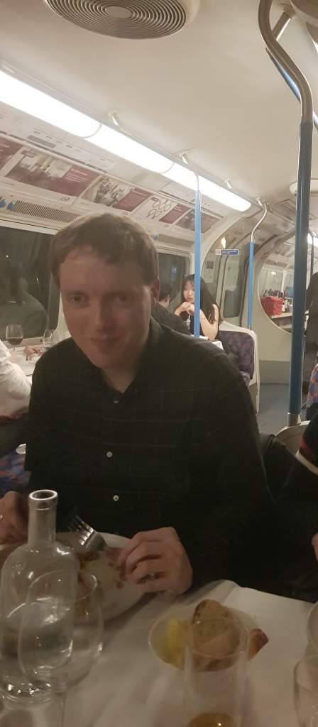 Glen eating dinner while seated in a old Victoria Line tube train carriage, with the original seating and blue poles.