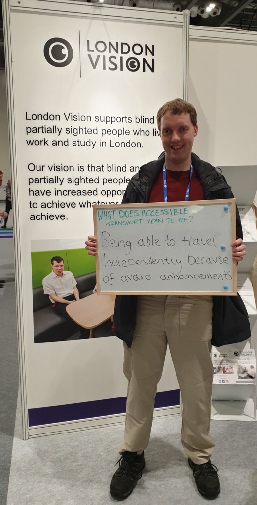 Standing in front of a tall London Vision banner, Glen is smiling and holding up a white board with handwritten text on it that says What does accessible transport mean to me? Being able to travel independently because of audio announcements.