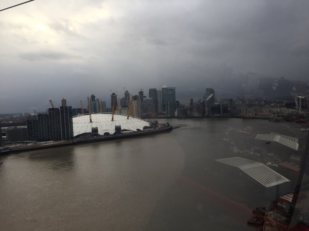 View from the Emirates Air Line cable car, showing the large white dome of the O2 Arena on the opposite side of the River Thames, with lots of skyscrapers in the background.