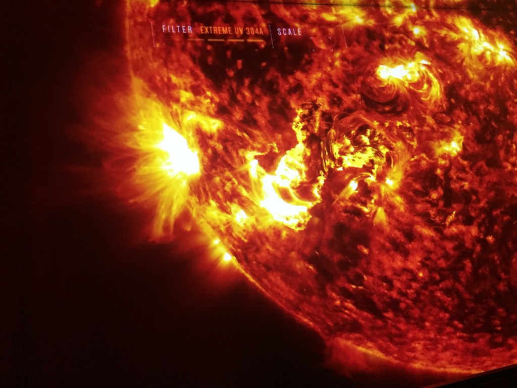 Close-up image of the sun using an extreme UV filter, which shows the surface as red with bright yellow light bursting out of it.