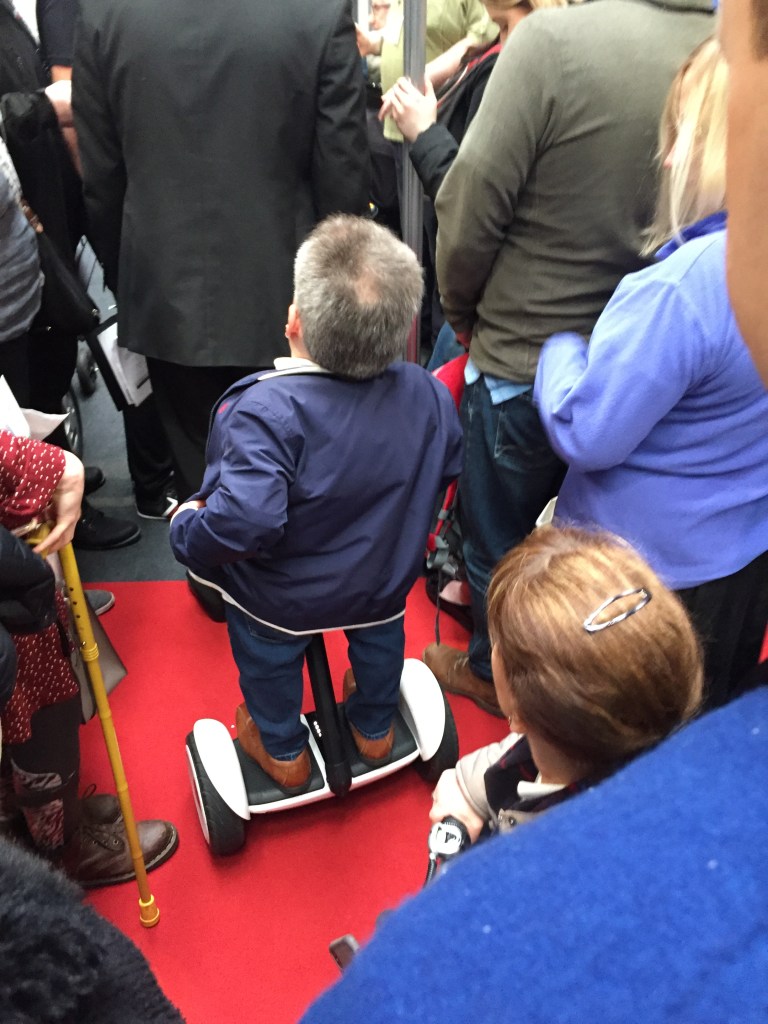 Warwick Davis standing on a small Segway machine, which is taking him through a large crowd of people.