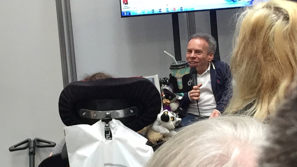 Warwick Davis sitting on a table and holding a microphone as he addresses the audience.