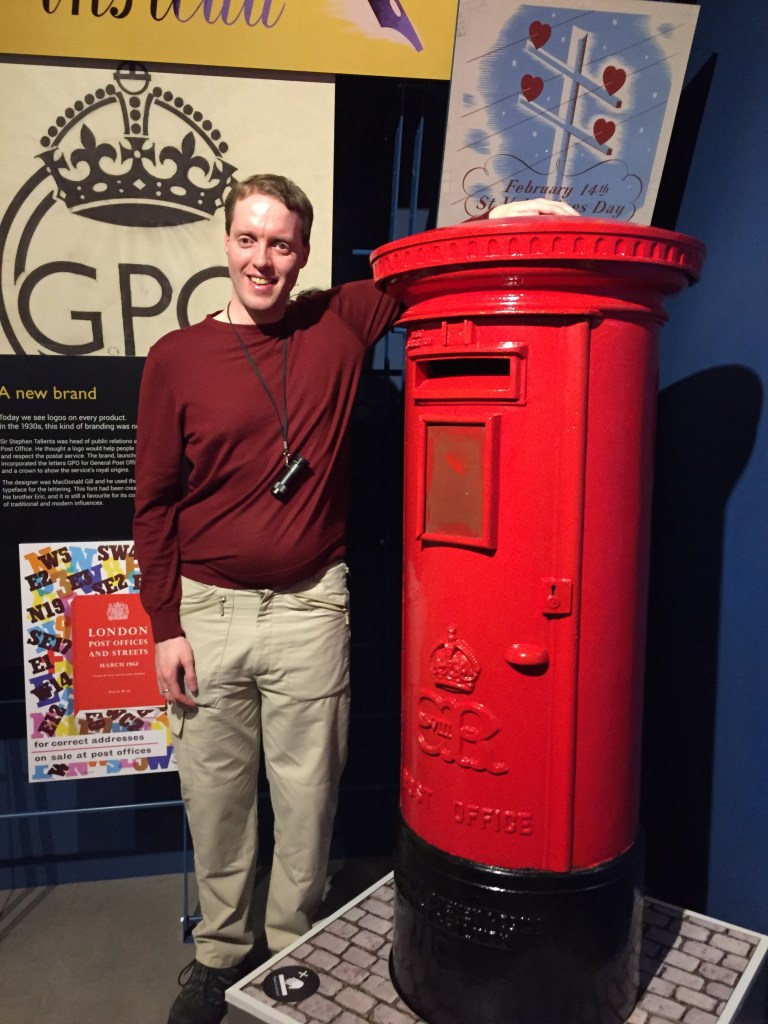 Glen smiling as he poses next to a red post box, resting his left arm on top of it. He's wearing a red top and light coloured cargo trousers, and has his monocular around his neck.