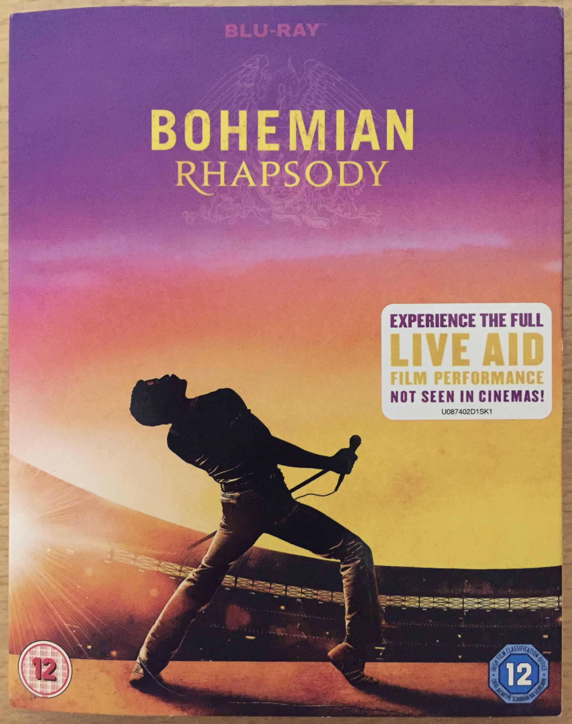 Blu-ray cover for the Bohemian Rhapsody movie. The image shows Freddie leaning back and holding the microphone stand across his front, while on stage during Live Aid in Wembley Stadium, under a multicoloured sky that ranges from purple to orange to yellow. A white sticker has text saying Experience the full Live Aid film performance not seen in cinemas.