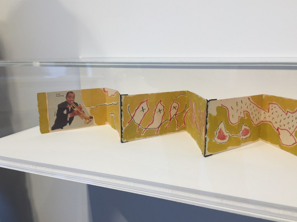 Opening section of a long, narrow book-like artwork, that's been unfolded in a zig-zagging manner along a shelf. The first page contains a photo of trumpeter Louis Armstrong, followed by a variety of patterns and colours across the various pages.