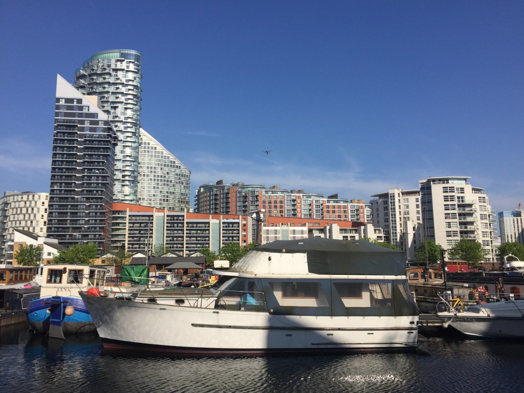 Boats parked in Poplar Dock Marina, with blocks of flats of varying heights and designs in the background, including a couple of tower blocks with diagonal sloping roofs, all under a clear blue sky.