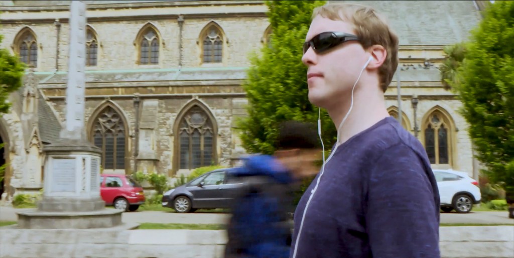 Wearing sunglasses and white Apple earphones, Glen looks around him as he walks past a large church.