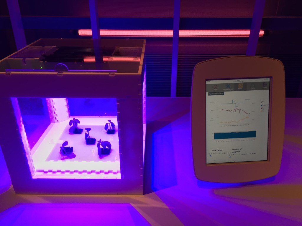 5 small plants under ultraviolet light in a large glass box, with a monitoring screen next to them showing measurements on line graphs.