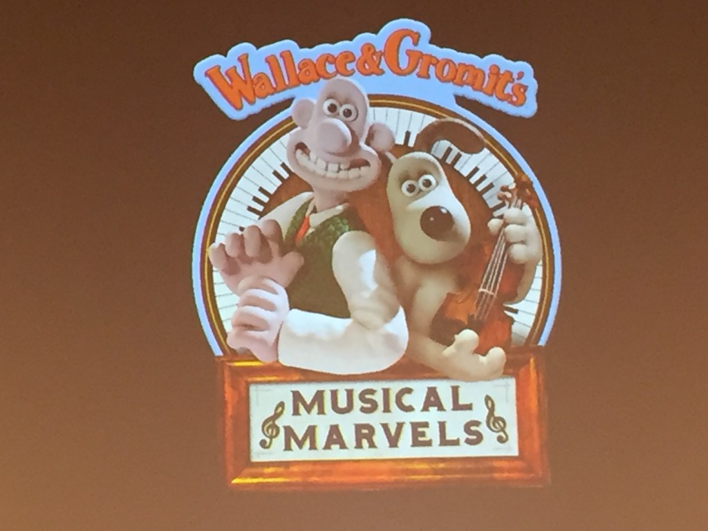 The characters of Wallace & Gromit standing next to each other. Wallace is smiling widely, while Gromit holding a violin. They are standing in front of a circle made of piano keys. Text reads Wallace & Gromit's Musical Marvels, with the character names above the circle, and the show title at the bottom.