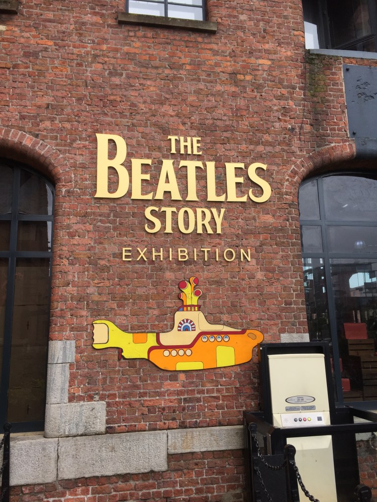 Sign for The Beatles Story Exhibition, written in gold letters on the brick wall of the museum. Below the lettering is a cartoon image of a yellow submarine.