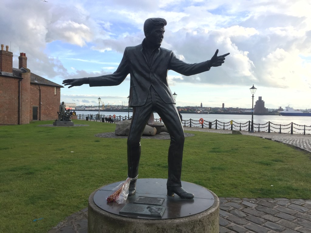 Life-size statue of Billy Fury, standing legs apart with one arm stretched out and pointing in front of him, and the other arm stretched out behind, as if he's in the middle of a dance move. The River Mersey is in the background.