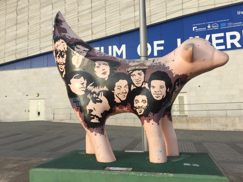 Statue of a lamb with a banana shaped tail. The head and legs are a plain light pink colour, but the body and tail are covered with black and white images of smiling faces against a black background.